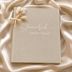 Op de afbeelding: Een beige, met linnen bedekt boek met het woord "Stammbuch" en "Familie Şimşek" in witte letters. Een klein bosje gedroogde bloemen en een lint zijn aan de rug van het boek bevestigd. Het boek ligt op een satijnen oppervlak.