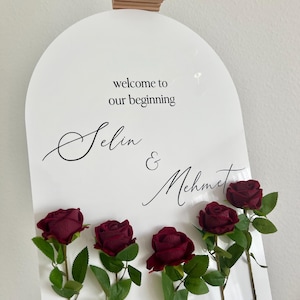 May include: An arched white sign featuring the black text "welcome to our beginning" followed by the names "Selin & Mehmet" in a script font. Five deep red artificial roses with green leaves are attached to the front of the sign.