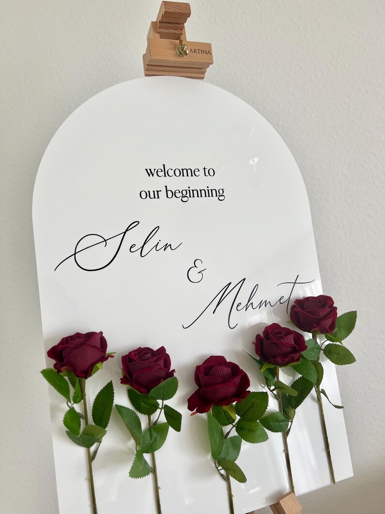 May include: An arched white sign featuring the black text "welcome to our beginning" followed by the names "Selin & Mehmet" in a script font. Five deep red artificial roses with green leaves are attached to the front of the sign.