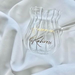 May include: A clear glass coffee pot with a fluted, vertical ribbed design and a scalloped rim. The glass features gold cursive text that reads "Hasan'in Kahvesi" on the front surface. It includes a single glass handle extending to the right.