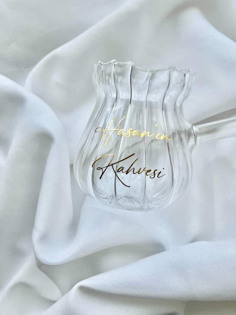 May include: A clear glass coffee pot with a fluted, vertical ribbed design and a scalloped rim. The glass features gold cursive text that reads "Hasan'in Kahvesi" on the front surface. It includes a single glass handle extending to the right.
