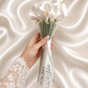 Könnte beinhalten: Ein Strauß weißer Calla-Lilien, eingewickelt in transparenten Stoff mit zartem Perlenbesatz. Ein langes weißes Band ist um die Stiele gebunden und mit dem gedruckten Text 06.06.2023 sowie den Namen Nicolas & Juleda in schwarzer Schreibschrift versehen.