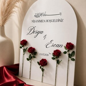 May include: An arched white sign featuring black text that reads "NIŞANIMIZA HOŞGELDINIZ", "Derya & Ercan", and "03.07.2026" with Arabic calligraphy at the top. Five individual red roses with green stems and leaves are attached to the front surface.