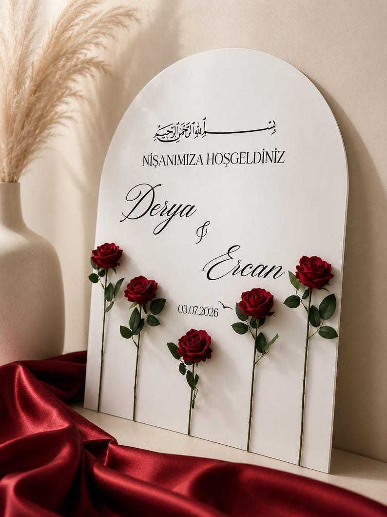 May include: An arched white sign featuring black text that reads "NIŞANIMIZA HOŞGELDINIZ", "Derya & Ercan", and "03.07.2026" with Arabic calligraphy at the top. Five individual red roses with green stems and leaves are attached to the front surface.