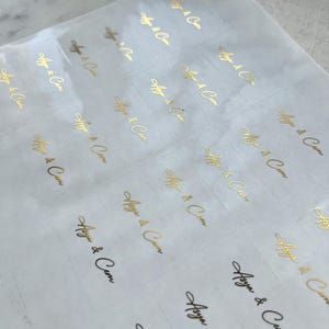 May include: A sheet of clear stickers with gold foil lettering. Each sticker has the text "Asya & Cem" in an elegant script. The stickers are arranged in neat rows and columns, ready for use.