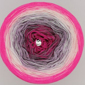 Könnte beinhalten: Eine Knäuel pinkfarbenes, weißes und graues Ombre-Garn mit einem silbernen Apfel-Anhänger.