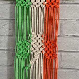Pode incluir: Uma tape&ccedil;aria de macram&eacute; verde, branca e laranja com uma barra de madeira no topo.