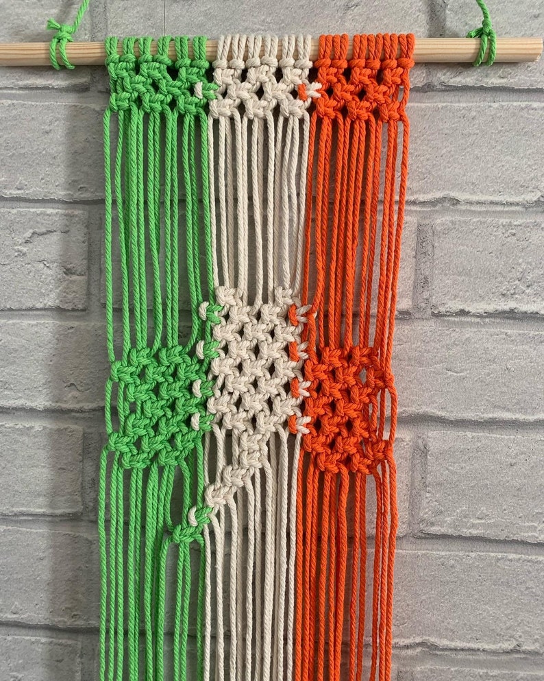 Pode incluir: Uma tape&ccedil;aria de macram&eacute; verde, branca e laranja com uma barra de madeira no topo.