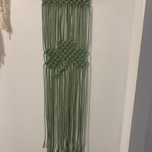 Pode incluir: Uma tape&ccedil;aria de macram&eacute; verde com um padr&atilde;o geom&eacute;trico. A tape&ccedil;aria &eacute; feita com cord&atilde;o de algod&atilde;o natural e est&aacute; presa a uma vareta de madeira.