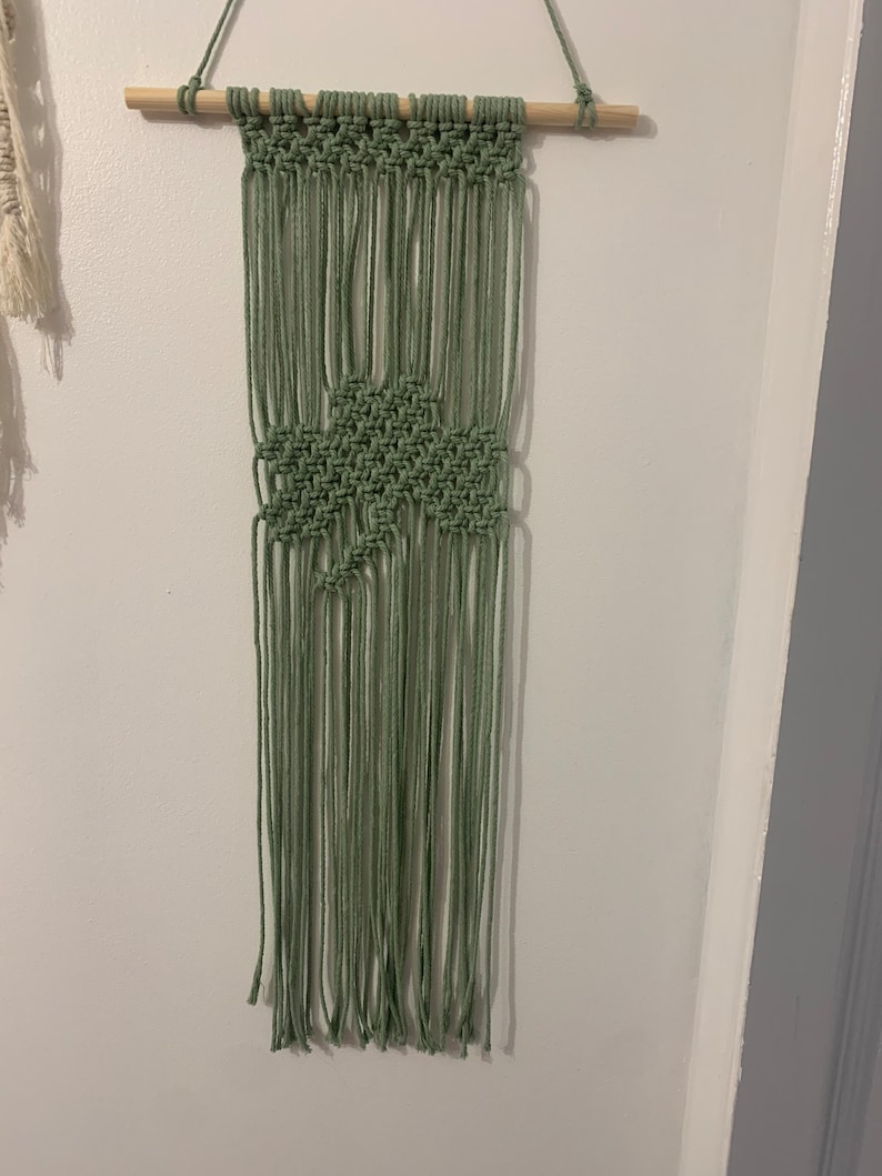 Pode incluir: Uma tape&ccedil;aria de macram&eacute; verde com um padr&atilde;o geom&eacute;trico. A tape&ccedil;aria &eacute; feita com cord&atilde;o de algod&atilde;o natural e est&aacute; presa a uma vareta de madeira.