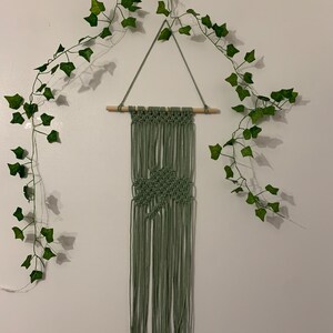 Pode incluir: Uma tape&ccedil;aria de macram&eacute; verde com um padr&atilde;o de diamante, pendurada em uma barra de madeira. A tape&ccedil;aria de parede &eacute; decorada com uma guirlanda de folhas verdes.