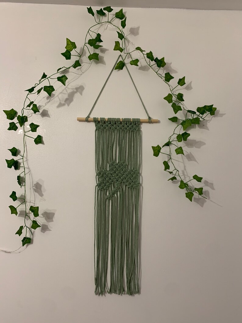 Pode incluir: Uma tape&ccedil;aria de macram&eacute; verde com um padr&atilde;o de diamante, pendurada em uma barra de madeira. A tape&ccedil;aria de parede &eacute; decorada com uma guirlanda de folhas verdes.