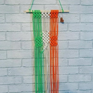 May include: A green, white and orange macrame wall hanging with a wooden dowel at the top. The hanging is made with knotted string and has a heart-shaped tag attached.