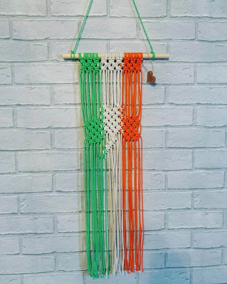 Pode incluir: Uma tape&ccedil;aria de macram&eacute; verde, branca e laranja com uma barra de madeira na parte superior. A tape&ccedil;aria &eacute; feita com corda atada e tem uma etiqueta em forma de cora&ccedil;&atilde;o.