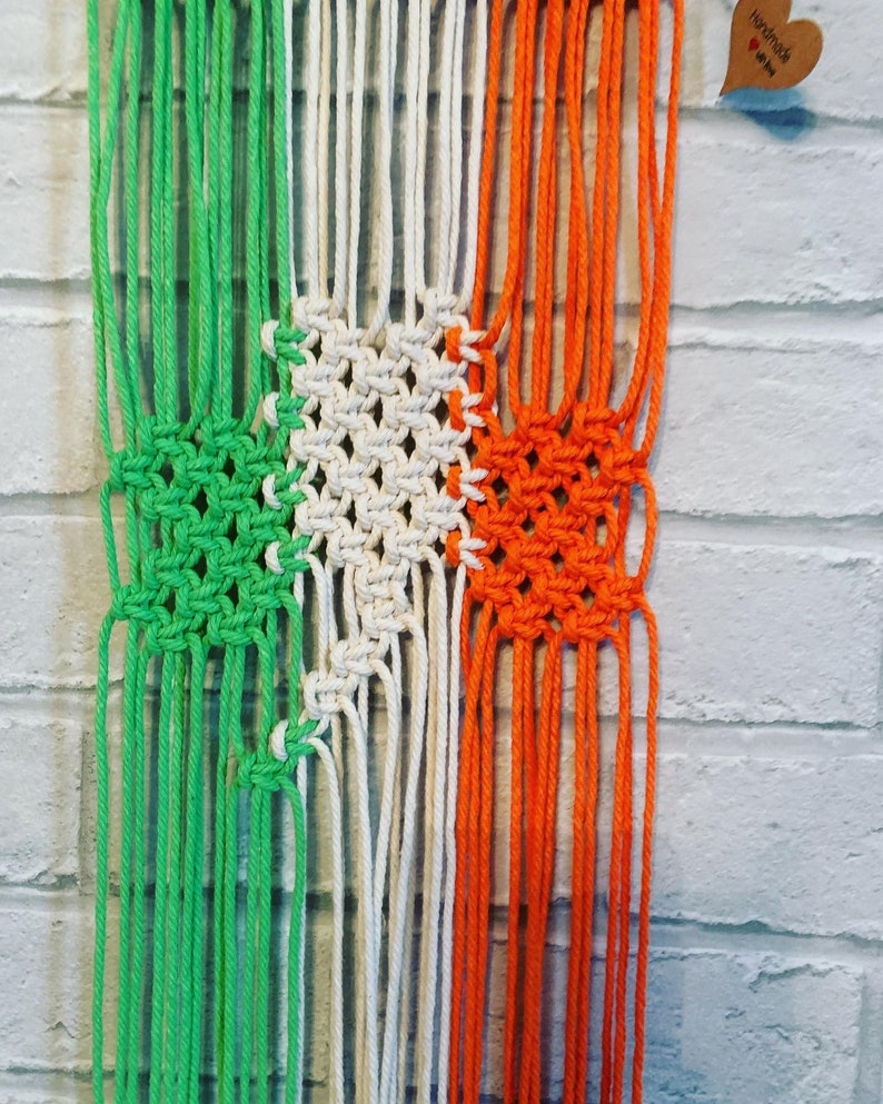 Pode incluir: Uma tape&ccedil;aria de macram&eacute; verde, branca e laranja com uma etiqueta "Handmade with love" anexada.