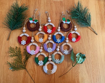 May include: A collection of 14 handmade Christmas ornaments. Each ornament is a wooden ring wrapped in colorful yarn with a white snowflake in the center. Two of the ornaments are brown and feature a green holly sprig with red berries.
