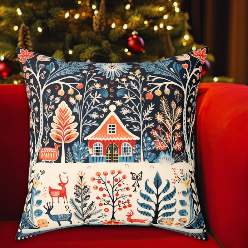 May include: Square cushion with a forest design featuring a red house, deer, and trees in blue, orange, and white on a dark blue backdrop. The cushion is placed on a red sofa, with a Christmas tree visible in the background, creating a festive atmosphere.