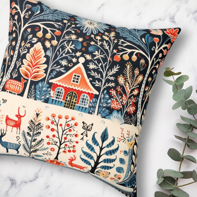 May include: Square cushion with a forest-themed design. The cushion has a navy blue backdrop with a cream-coloured band. The illustration includes a red house, deer, trees, and floral motifs in orange, blue, and cream hues. The cushion is square.