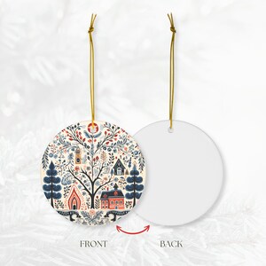 May include: A round ceramic Christmas ornament with a folk art design. The front features a tree with houses, flowers, and other details in red, blue, and beige. The back is plain white. A gold string is attached for hanging.