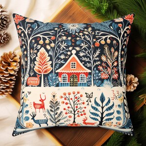 May include: Square cushion with a forest-themed design. The pattern includes a red house, deer, trees, and floral motifs in navy, red, orange, and blue. The cushion is displayed on a wooden tray, with pine needles and pine cones.