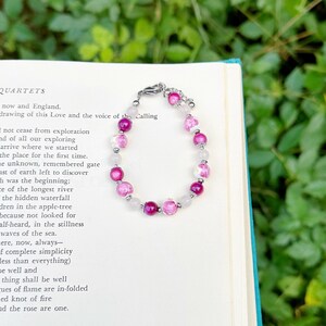 May include: A beaded bracelet featuring a mix of pink and white beads, interspersed with clear, round beads. The bracelet is made with a silver-colored chain and clasp. The bracelet is resting on an open book with text.