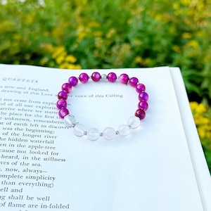 May include: A beaded bracelet featuring alternating pink and clear gemstone beads. The bracelet is resting on a book with text, set against a blurred background of green foliage and yellow flowers.