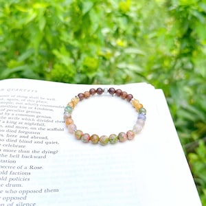 May include: A beaded bracelet with a variety of colorful gemstones, including brown, green, and yellow, rests on an open book. The bracelet's beads are round and vary in size. The background is a blurred view of green foliage.