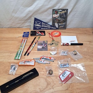 May include: A collection of Dale Earnhardt Jr. racing memorabilia including a Daytona International Speedway pennant, a collectable knife, pencils, pens, patches, pins, and stickers featuring the Club E Jr. logo and race car imagery.