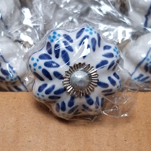 May include: A white ceramic cabinet knob with a scalloped, flower-like shape, decorated with a blue leaf and dot pattern. The center features a silver-toned metal rosette with a small, smooth, circular silver-toned accent in the middle.