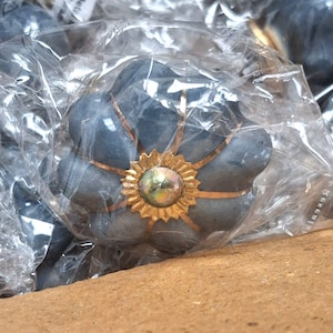 May include: A flower-shaped cabinet knob with a matte dark gray finish, featuring thin gold-colored metal accents radiating from a central circular gold-toned floral embellishment with a small iridescent center, encased in clear plastic packaging.
