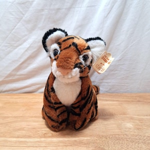 May include: A plush tiger toy with orange and black stripes, a white chest, and a white muzzle. The tiger has large dark eyes, rounded ears with white inner fur, and a tan tag attached to its ear that reads SEMO CUDDLY TOYS.