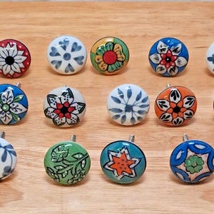 May include: A collection of ceramic cabinet knobs with various floral and geometric designs. The knobs feature a range of colors, including blue, red, green, orange, and white, and are arranged on a wooden surface.