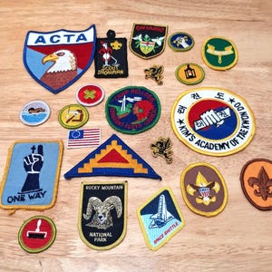 May include: A collection of various embroidered fabric patches featuring diverse shapes, colours, and designs, including an eagle, a space shuttle, a ram, a fleur-de-lis, and text such as ACTA, Scout Showman, Ontario, Kim's Academy of Tae Kwon Do, and Rocky Mountain.