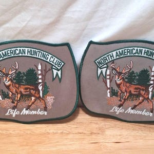 May include: Two identical taupe-colored patches featuring an embroidered deer standing in a forest scene. Each patch displays the text North American Hunting Club in a banner above the deer and the words Life Member in script below the deer.