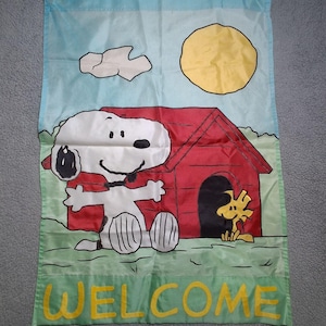May include: A rectangular garden flag featuring a light blue sky with a yellow sun and a white cloud. A white Snoopy dog sits in front of a red doghouse with a yellow Woodstock bird nearby. The word WELCOME is printed in yellow at the bottom.