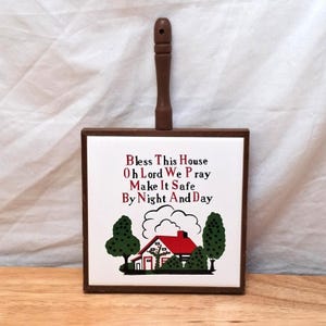 May include: A square wooden wall plaque with a white ceramic tile insert featuring a red-roofed house illustration, green trees, and the text: Bless This House Oh Lord We Pray Make It Safe By Night And Day. The plaque has a brown wooden handle.