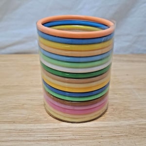 May include: A stack of colorful plastic bangles in a clear plastic wrap. The bangles are in various colors, including orange, blue, yellow, green, and pink. The bangles are stacked in a cylindrical shape. The background is a light brown wooden surface.