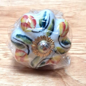 May include: A white ceramic cabinet knob with a hand-painted floral pattern featuring red and yellow tulips and green leaves. The knob has a scalloped edge and a central metallic gold-tone flower-shaped decorative plate, all wrapped in clear plastic.