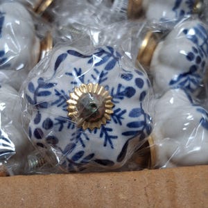 May include: A white ceramic cabinet knob with a scalloped edge and a blue floral pattern. The center features a gold-toned metal rosette and a screw attachment. The knob is individually wrapped in clear plastic and sits inside a cardboard box.