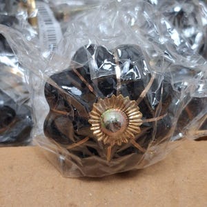 May include: A black, flower-shaped cabinet knob featuring a gold-toned metal center with a sunburst design and thin gold-toned lines radiating outward across the petals, all encased in a clear, crinkled plastic protective packaging.