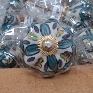 May include: A round ceramic drawer knob featuring a white base with a painted pattern of teal, dark blue, and yellow shapes. The center of the knob has a gold-colored metal sunburst-shaped decorative plate with a smooth, reflective silver-toned dome.
