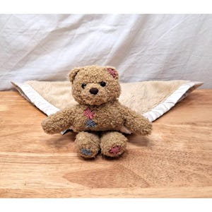 May include: A tan teddy bear with curly fur, featuring small red and blue checkered patches on its left ear, chest, and feet. The bear is attached to a soft, beige, triangular-shaped security blanket with a white satin trim border.