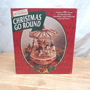 May include: A red cardboard box for a Mr. Christmas Christmas Go Round carousel. The box features a graphic of a musical carousel with horses and riders, text reading "Listen to 25 of your all time favorite Christmas songs while the carousel turns."