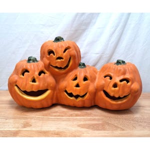 Peut inclure: Un groupe décoratif de citrouilles en céramique orange représentant quatre jack-o'-lanterns aux visages souriants. Chaque citrouille présente des yeux, un nez et une large bouche découpés sans dents. Finition orange mat avec tiges vertes.