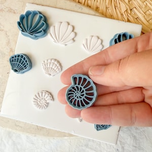 May include: A collection of seashell-shaped polymer clay cutters in various designs and colors, including blue and white. The cutters are arranged on a white surface, with one held in a hand. The theme is coastal and nautical.