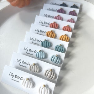 May include: A collection of handmade pumpkin-shaped stud earrings in various colors, including white, gray, blue, orange, and purple. Each earring has a gold-colored stem. The earrings are displayed on white cards with the text "Lily Rose Co Handmade".