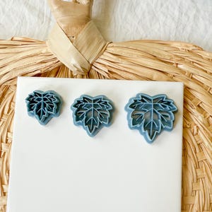 May include: Three blue leaf-shaped cookie cutters of varying sizes are arranged on a white surface. The cutters are made of plastic and have detailed leaf vein patterns. The background features a woven straw-like material.