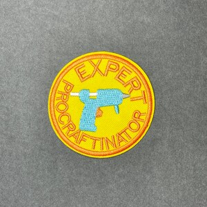 May include: A yellow embroidered patch with the words "EXPERT PROCRASTINATOR" in orange around the edge. A blue glue gun is in the center. The patch is circular.