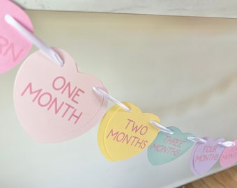 Könnte beinhalten: Ein rosa, gelber, grüner, lila und blauer Herz-Baby-Meilenstein-Banner mit den Worten "Geboren", "Ein Monat", "Zwei Monate", "Drei Monate", "Vier Monate" und "Fünf Monate", die auf jedes Herz gedruckt sind.