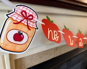 Può includere: Un banner decorativo con un barattolo di marmellata e ritagli a forma di mela rossa con numeri e lettere. Il barattolo ha un fiocco rosso e un coperchio a quadretti. Lo striscione è infilato su un nastro bianco.